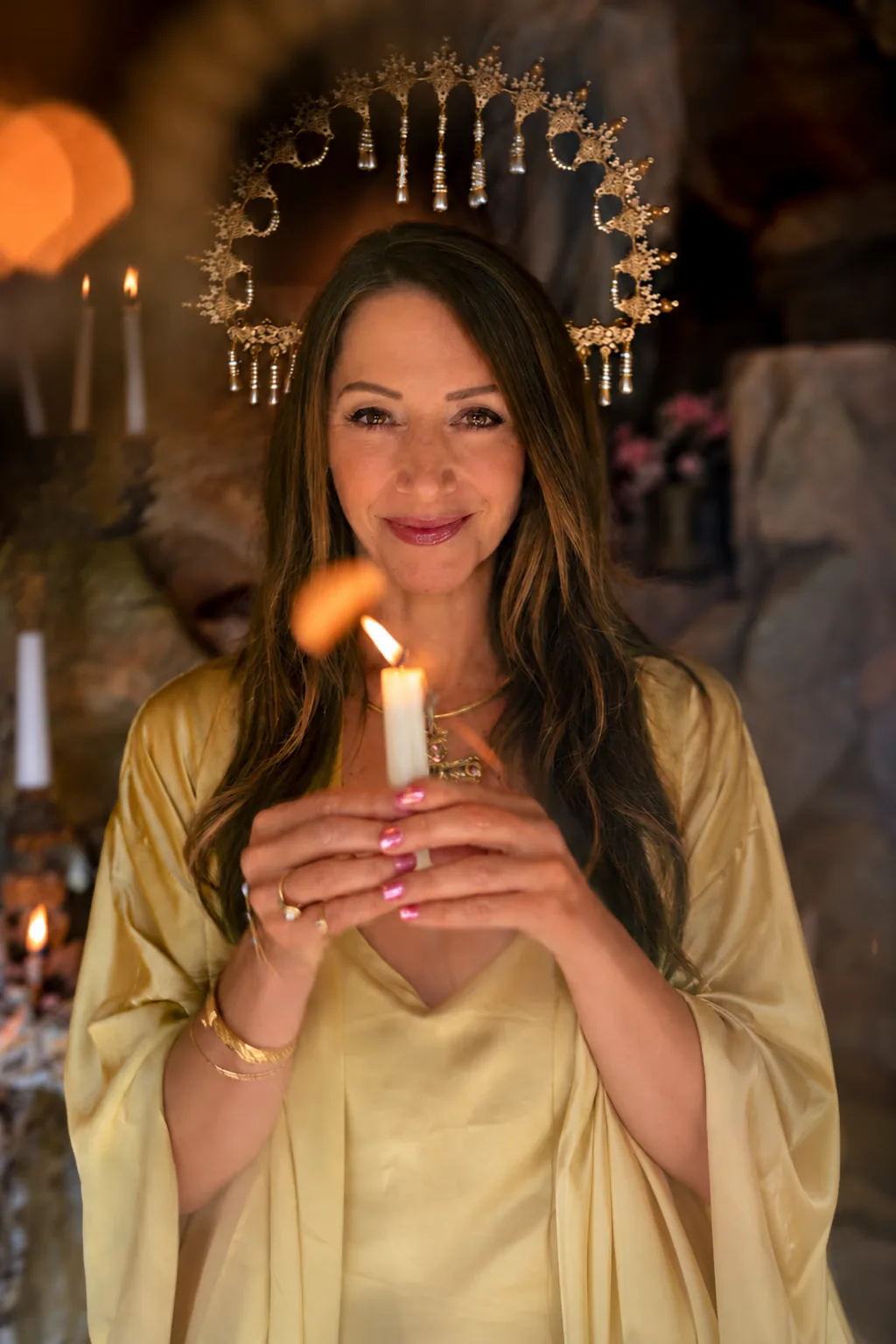 Christine Mira Rose in a candlelit grotto, wearing a gold halo crown and a pale gold silk robe, smiling as she holds a lit candle