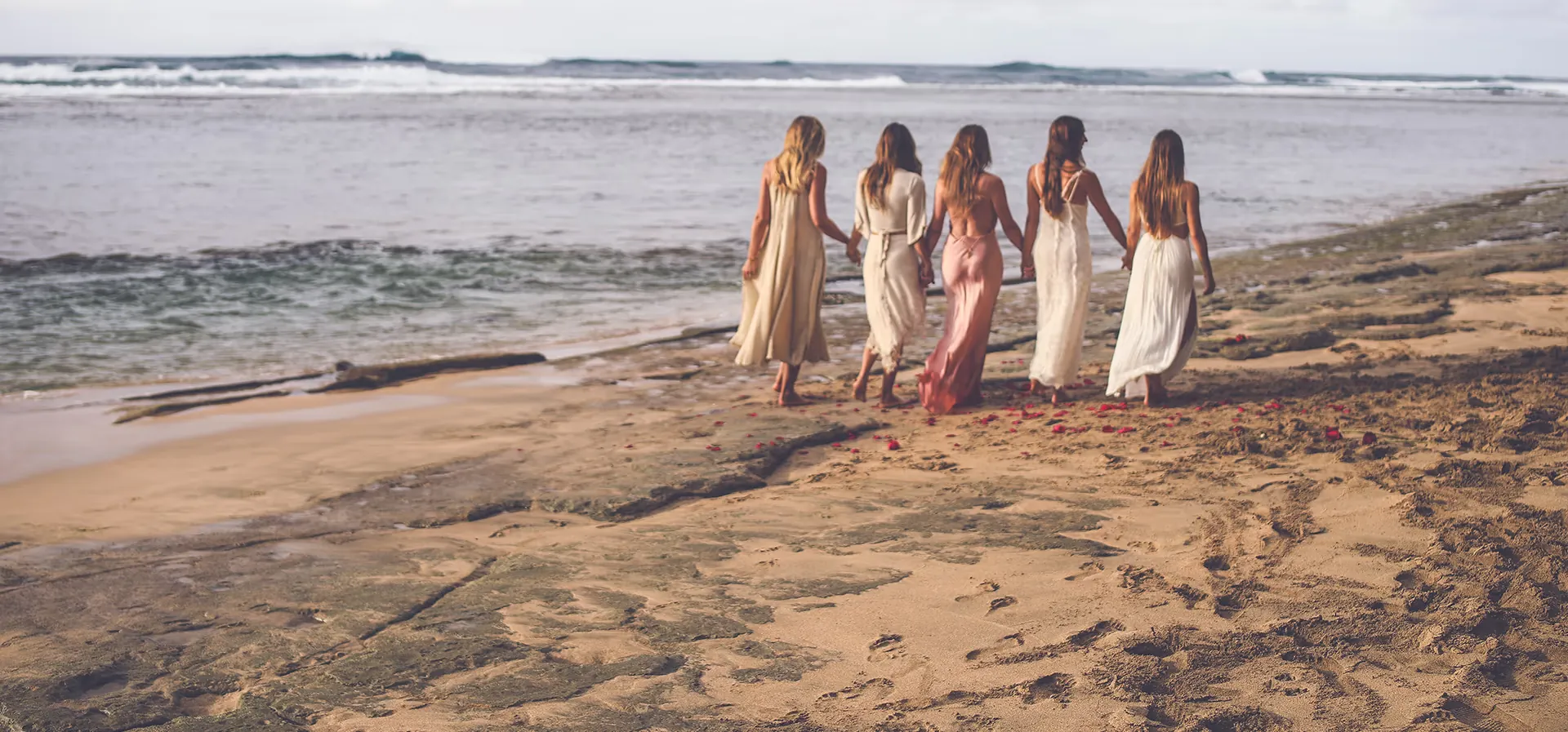 Five women in long cream, white and rose dresses walking hand in hand along the water&rsquo;s edge of a beach, seen from behind, with rose petals scattered on the sand behind them